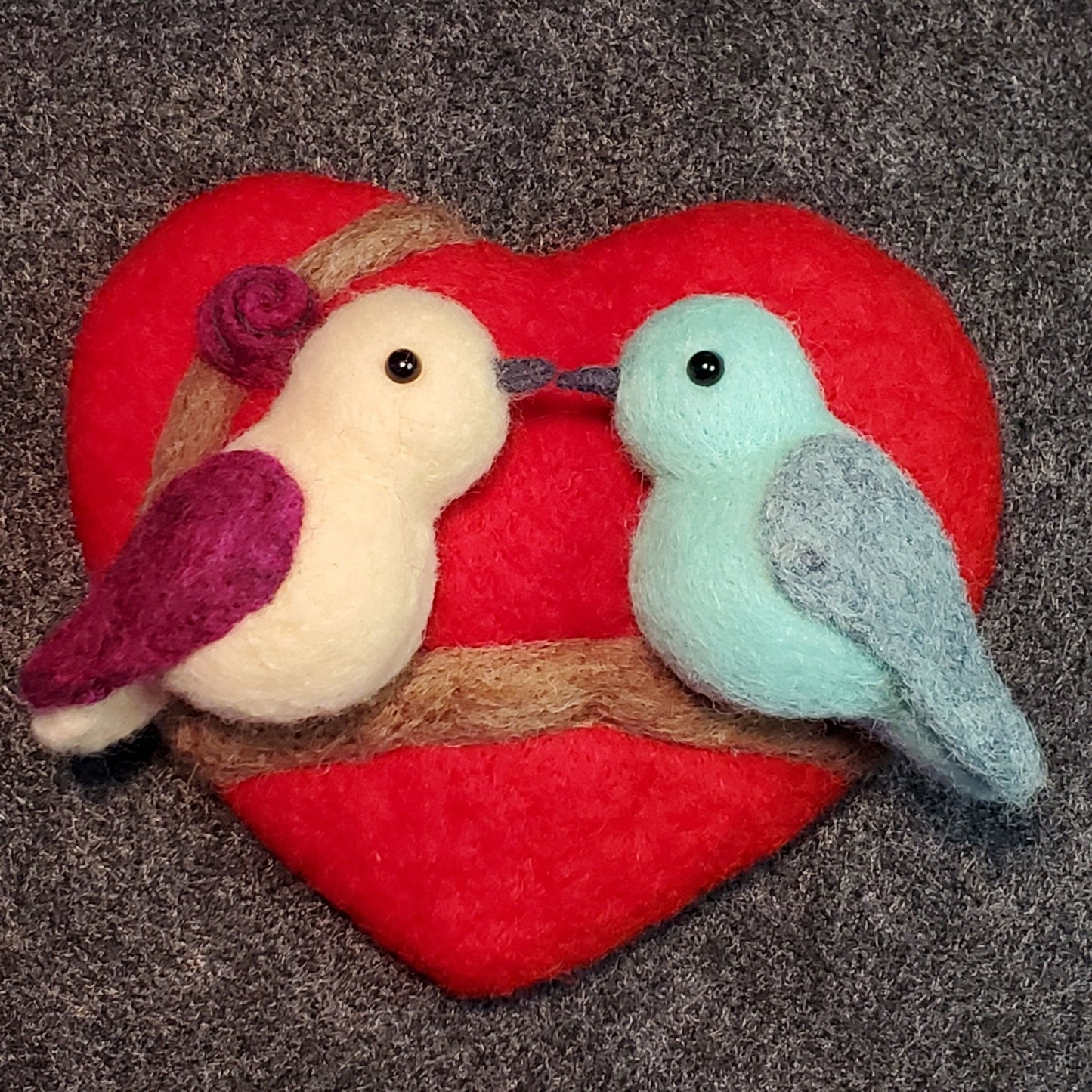 A needle-felted craft featuring two lovebirds perched on a heart-shaped wool pad, with a red background.